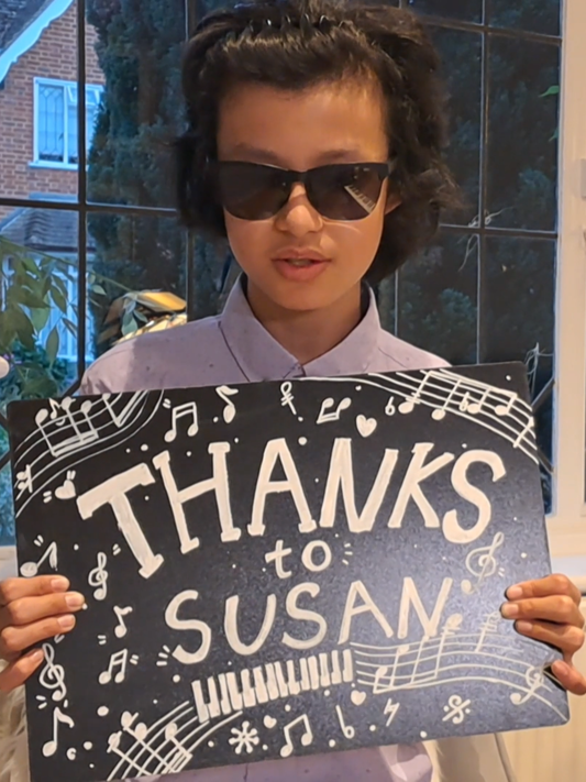 Chapman, sitting at his piano in his home, holding a sign that reads 'Thanks to Susan', surrounded by drawn musical notes