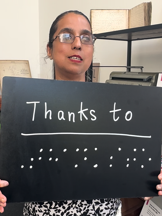Amrit standing in an office with shelves around her, holding a sign that reads 'Thanks to...' followed by braille writing.