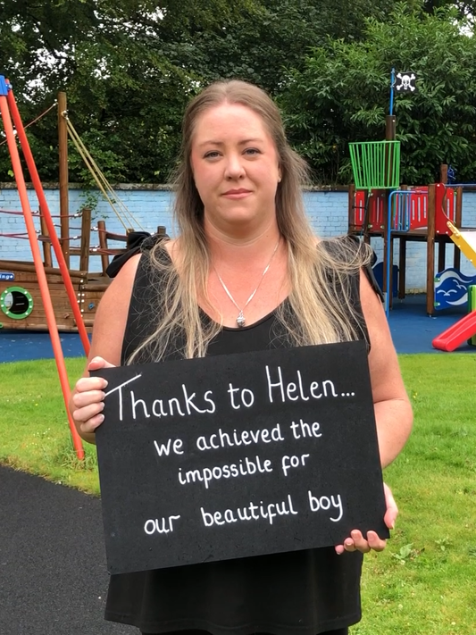 Cassy in the playground of Rachel House, holding a sign that reads 'Thanks to Helen, we achieved the impossible for our beautiful boy'